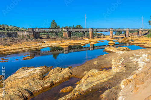 Reflejos de un puente de hierro en un río que fluye entre piedras amarillas