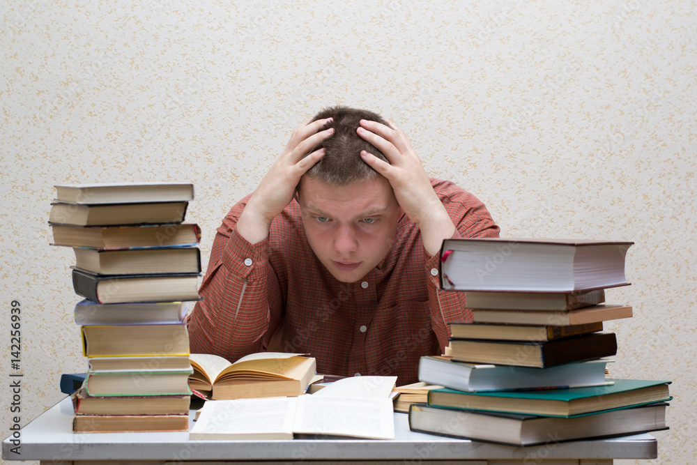 Confused guy sitting at a table with a pile of books Confused guy sitting at a table with a pile of books