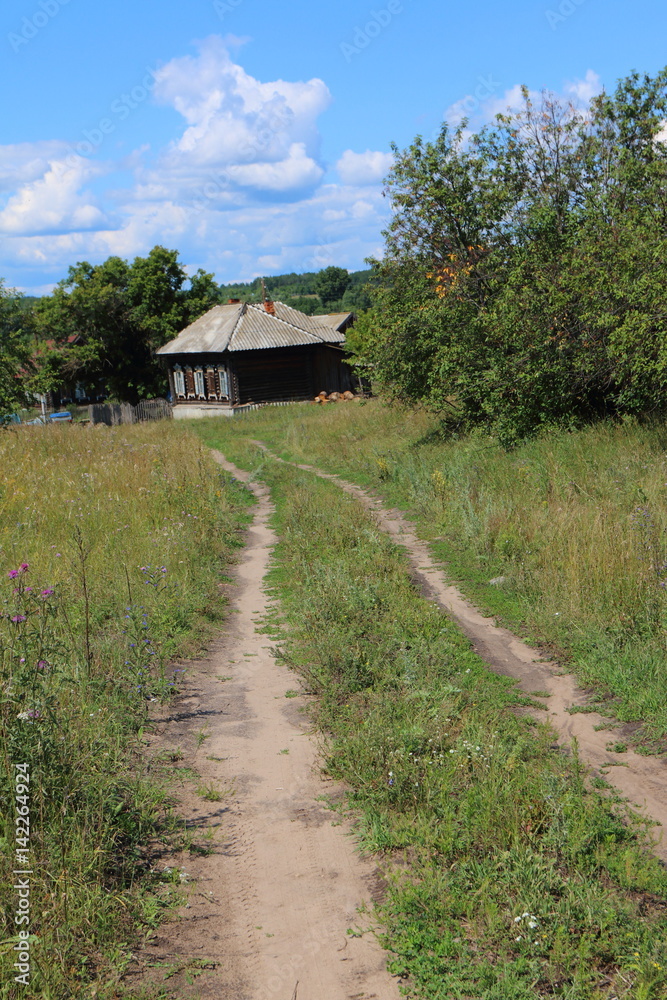 Dirt road leading to a lonely wooden house in the distant Russian village against a blue cloudy sky Dirt road leading to a lonely wooden house in the distant Russian village against a blue cloudy sky