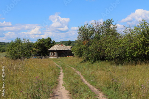 Dirt road leading to a lonely wooden house in the distant Russian village against a blue cloudy sky