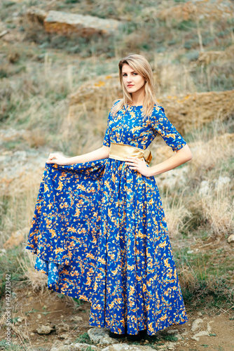 Beautiful Attractive Blond Caucasian Girl Model Stands Mountains and Sea Background in Long Blue Dress with Gold Yellow Pattern