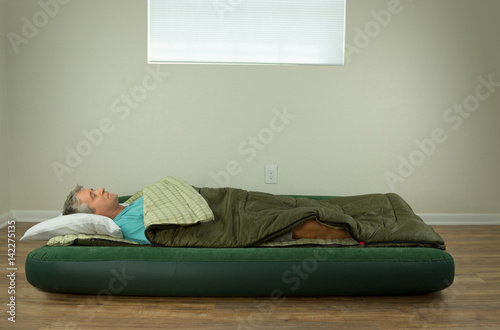 Man comfortably sleeping on blow up air mattress bed in sleeping bag with clean walls and window in the background.