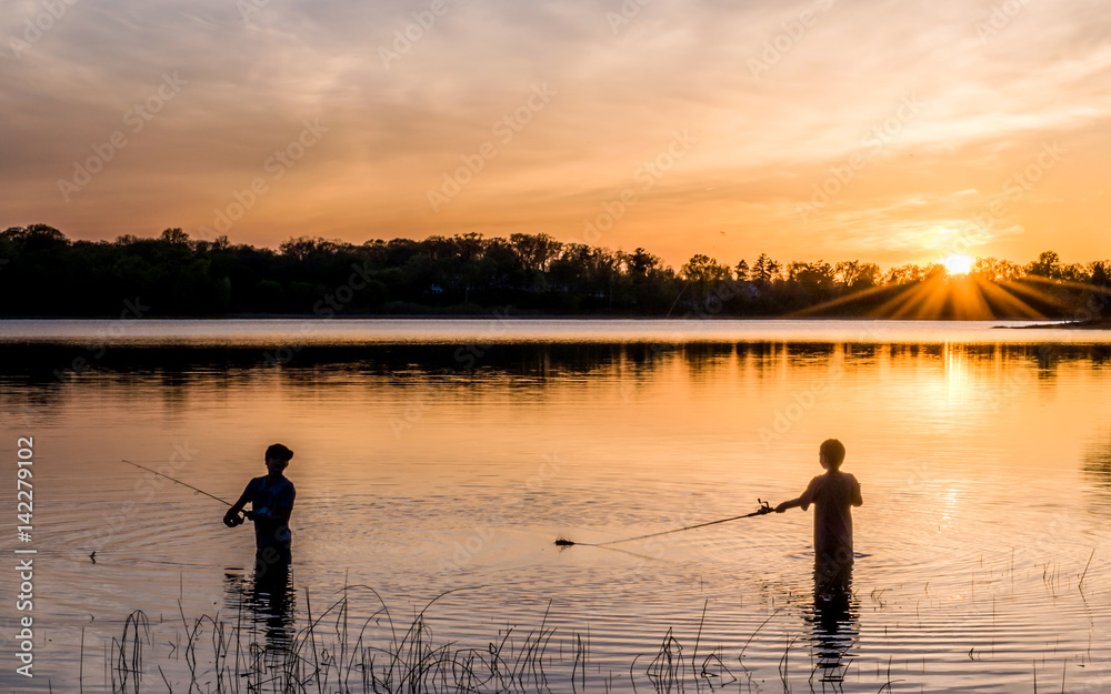 Two young kids are fishing under sunset of a lake