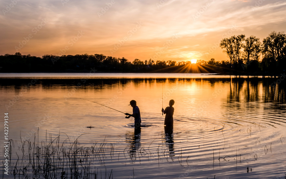 Two young kids are fishing under sunset of a lake