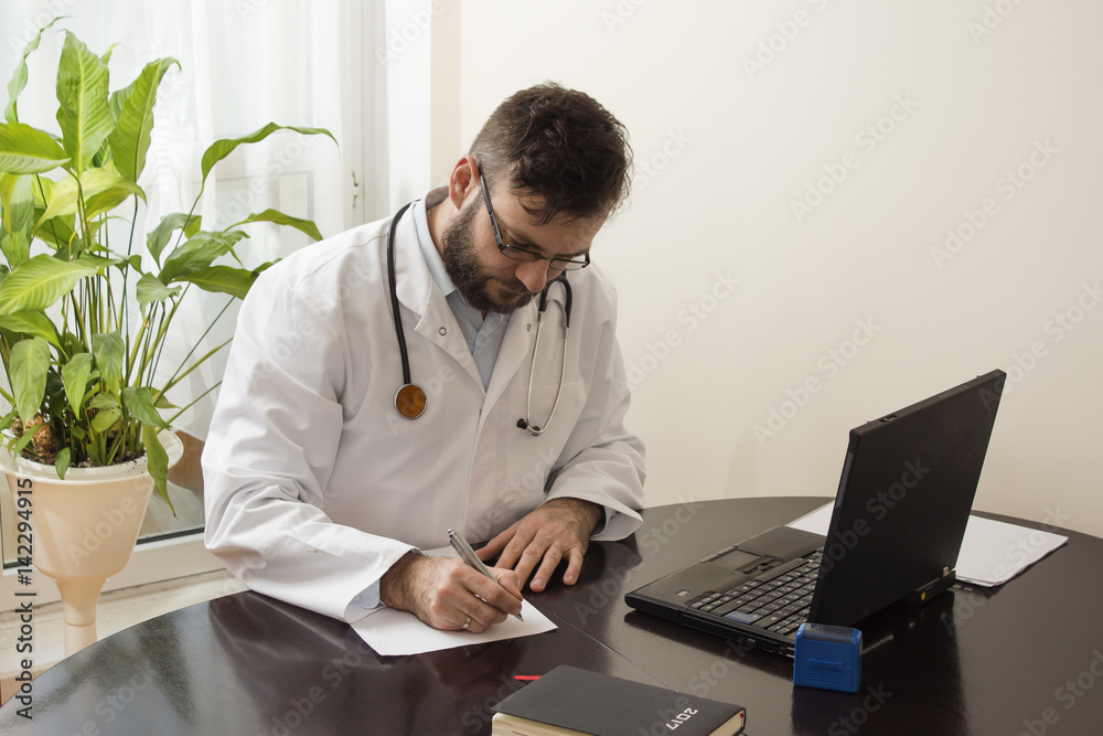 Lekarz w białym kitlu w gabinecie lekarskim siedzi przy stole i wypełnia dokumenty. Lekarz podczas pracy.  - obrazy, fototapety, plakaty 