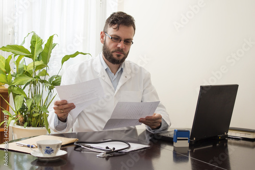  Lekarz w gabinecie siedzi przy biurku i przegląda dokumentację medyczną. Lekarz podczas pracy. 