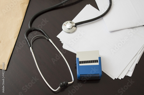 Biurko lekarza. Stetoskop, pieczątka i dokumenty leżą na ciemnym stole. Gabinet lekarski. Stetoskop, pieczątka, długopis i historia choroby. Atrybuty lekarza leżą na stole.