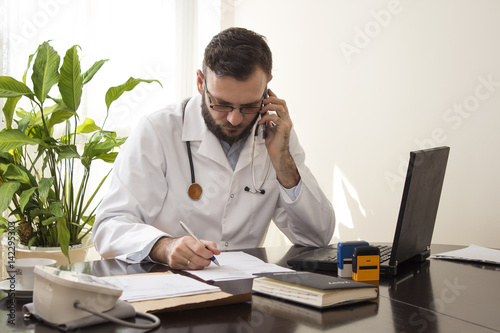 Lekarz w gabinecie lekarskim siedzi przy biurku z telefonem komórkowym w dłoni. Lekarz siedzi przy biurku, robi notatki rozmawiając przez telefon. Telefoniczna konsultacja lekarska.