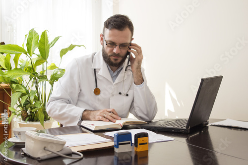 Lekarz w gabinecie lekarskim rozmawia przez telefon i sprawdza wolne terminy w kalendarzu. Lekarz w gabinecie lekarskim siedzi przy biurku z telefonem komórkowym w dłoni photo
