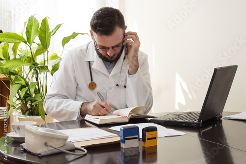 Lekarz w gabinecie lekarskim siedzi przy biurku z telefonem komórkowym w dłoni. Lekarz podczas rozmowy telefonicznej zapisuje termin spotkania w kalendarzu.