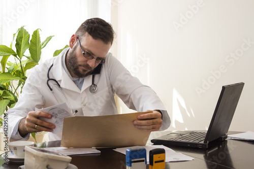 Lekarz siedzi przy biurku. Rozmawia przez telefon przeglądając dokumenty medyczne pacjenta