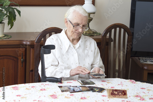 Stara kobieta siedzi w salonie przy stole i przegląda stare fotografie. Babcia staruszka ogląda stare zdjęcia siedząc przy stole.