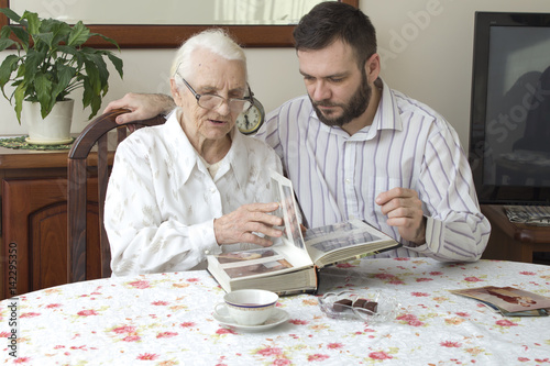 Staruszka z wnuczkiem siedzą przy stole w salonie i oglądają stare zdjęcia. Babcia z wnuczkiem wspominają dawne czasy oglądając album ze zdjęciami. Wnuczek pokazuje babci album ze zdjęciami. Spotkanie