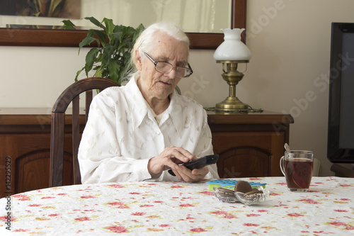 Old woman at the table with remote control for the TV. The old woman turns on the TV remote control sitting at the table. The old woman in glasses sitting at the table and looking for 