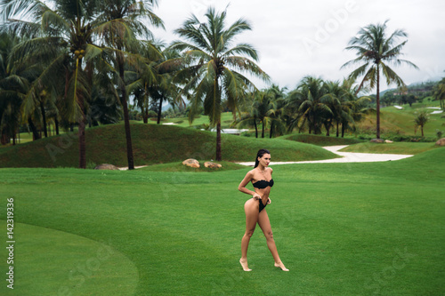 Young sexy and beautiful brunette fitness girl in black swimsuit posing turning booty shows ass at the green grass of Golf club. Sexy model portrait with perfect body. Concept of summer holiday