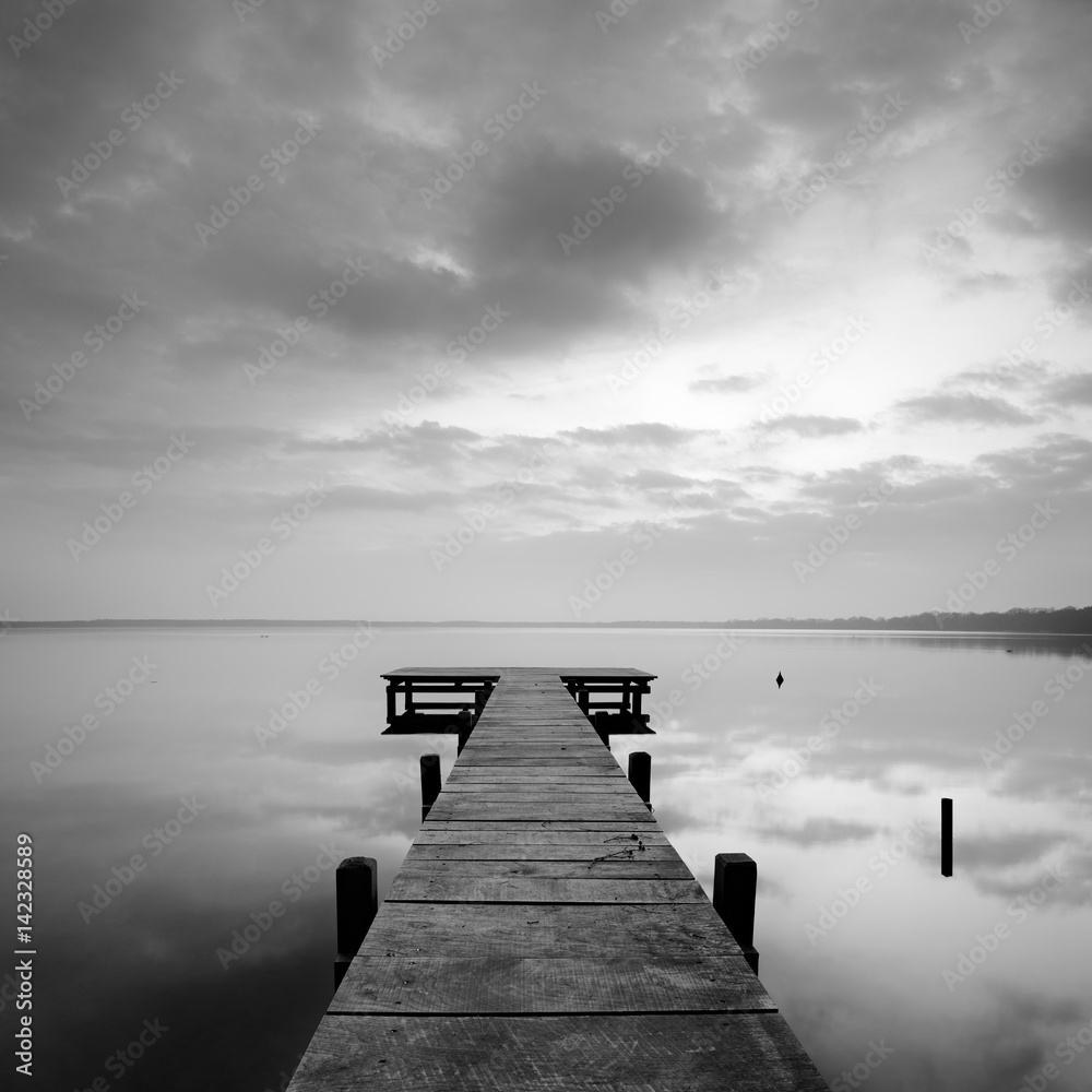 Fototapeta premium Stiller See mit Steg bei Sonnenaufgang, wolkiger Himmel, schwarz-weiß