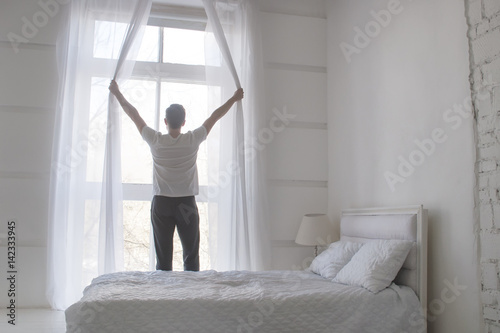 Young man opening curtains to welcomw morning and light  rear view  white