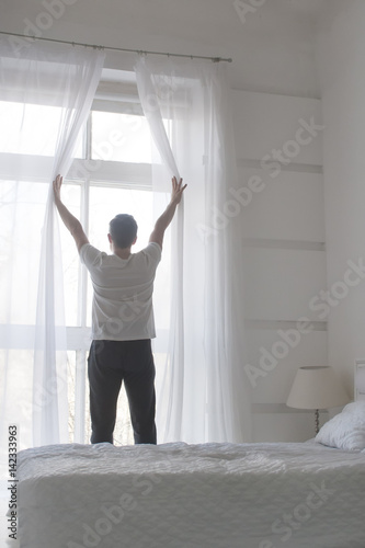 Young man opening curtains to welcomw morning and light, rear view, white