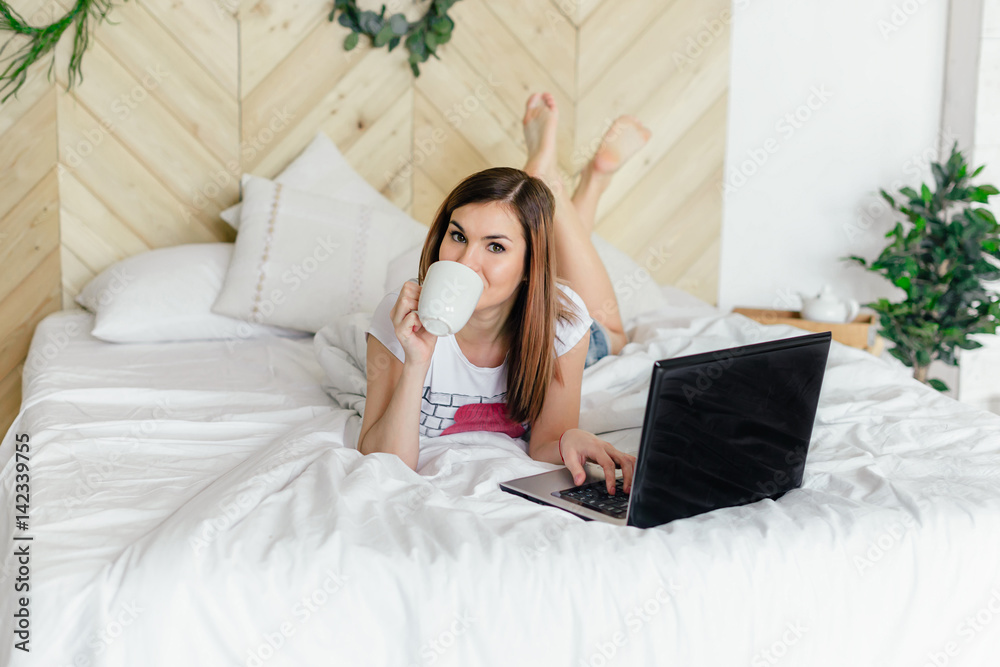 Obraz premium The girl in the bedroom on the bed under white blanket with a laptop and coffee in hand. Business girl. The morning starts with news on the Internet. Lifestyle people concept.
