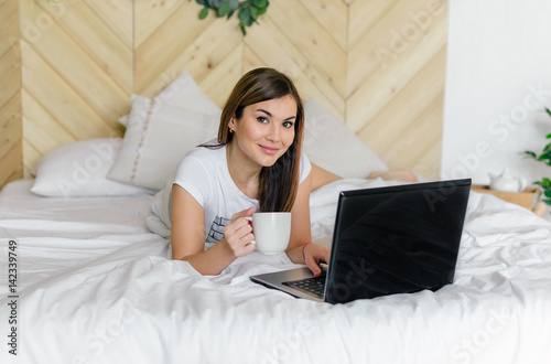 The girl in the bedroom on the bed under white blanket with a laptop and coffee in hand. Business girl. The morning starts with news on the Internet. Lifestyle people concept.