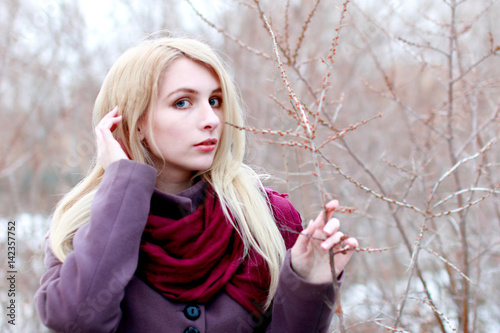 Beautiful blonde with bush branch looks at us, hand to head, light background.