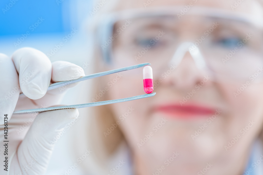 senior scientist in protective gloves and glasses holding pill with tweezers senior scientist in protective gloves and glasses holding pill with tweezers