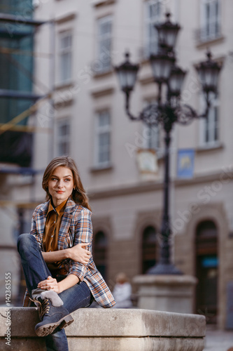 Fashion girl is sitting outdoors wearing blue jeans,brown checkered jacket and holding a brown handbag, urban city