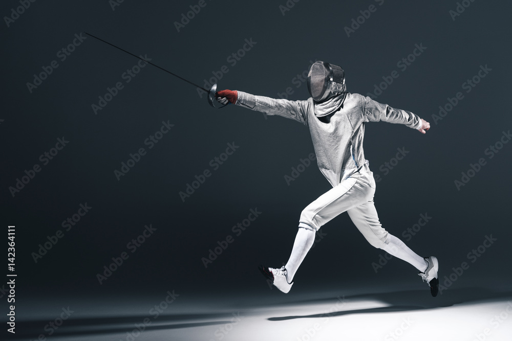 Professional fencer in fencing mask with rapier jumping on grey Professional fencer in fencing mask with rapier jumping on grey