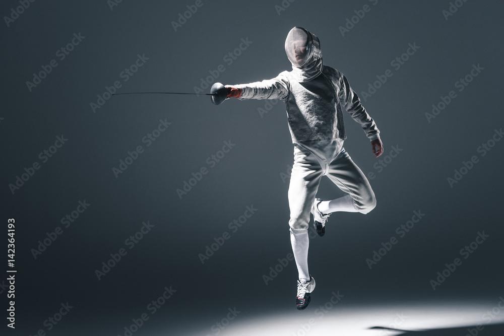 Professional fencer in fencing mask with rapier jumping on grey Professional fencer in fencing mask with rapier jumping on grey