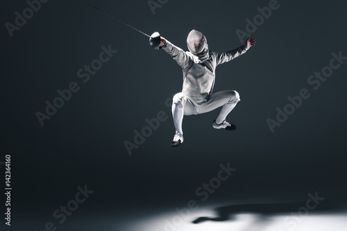 Professional fencer in fencing mask with rapier jumping on grey photo