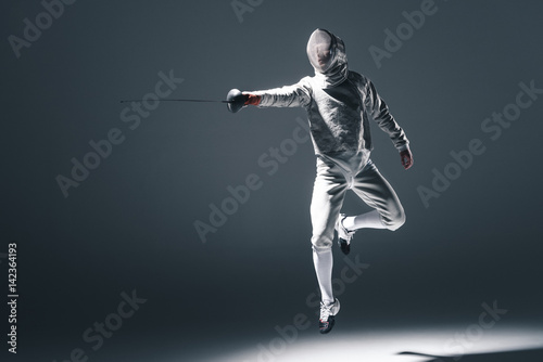 Professional fencer in fencing mask with rapier jumping on grey