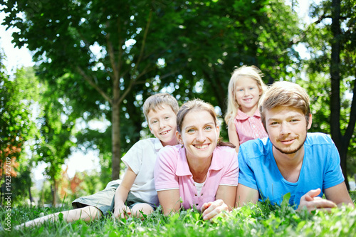young happy family at noon in the park on the grass. Two young parents and children, boy and girl, lies on the grass and smiling looking at the camera. © vitaliymateha