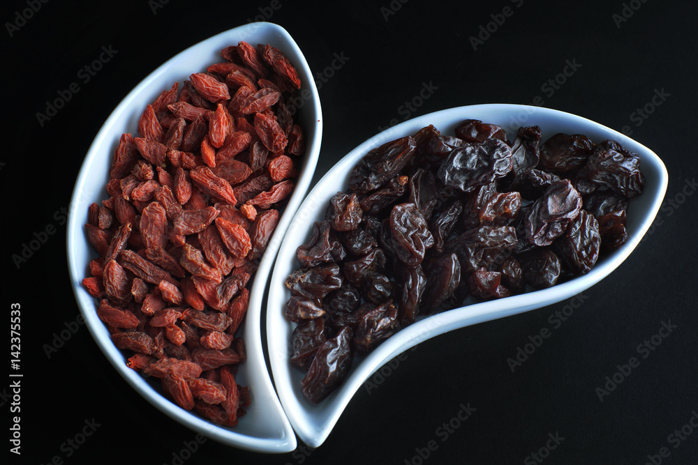 Dried goji berries and dark grapes in white bowls on a black background Dried goji berries and dark grapes in white bowls on a black background