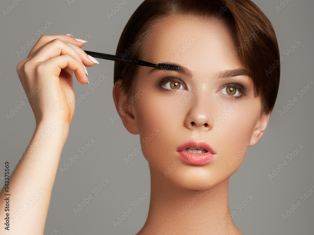 Beautiful young woman applying foundation on her face with a make up brush isolated on gray background