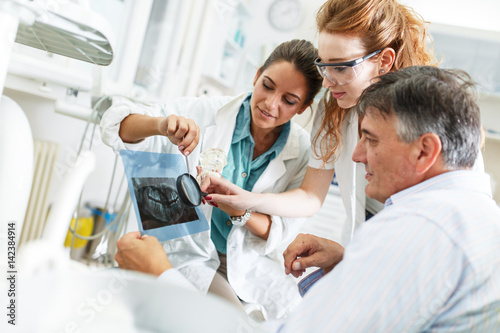 Dentist and her female assistant in dental office talking with senior patient and preparing for treatment.Examining x-ray image.