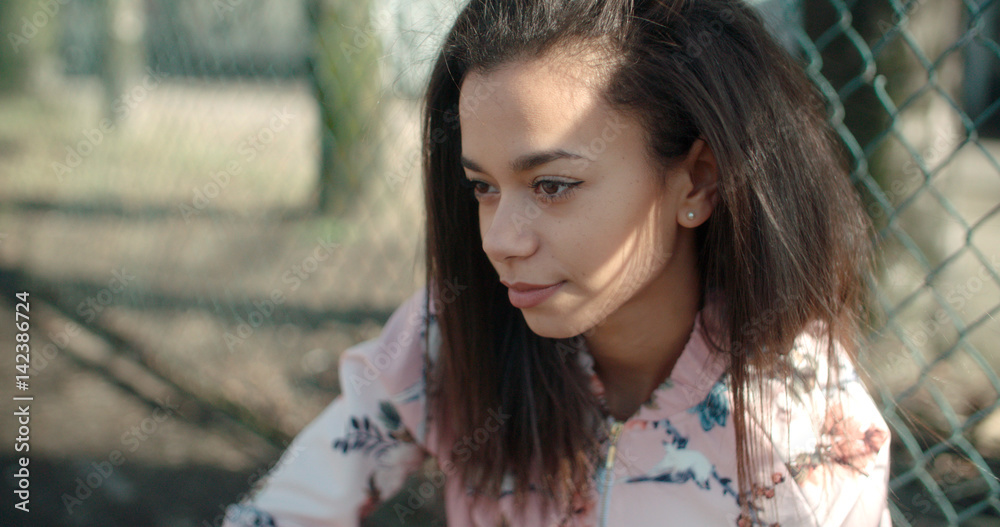 Portrait of a young African American woman wearing bomber jacket posing over metal fence, outdoors.