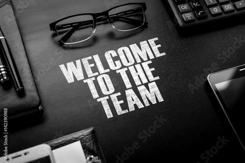 Black chalkboard with business accessories (notepad, magnifying glass, fountain pen, tablet, phone, glasses and calculator) and text WELCOME TO THE TEAM. Top view.