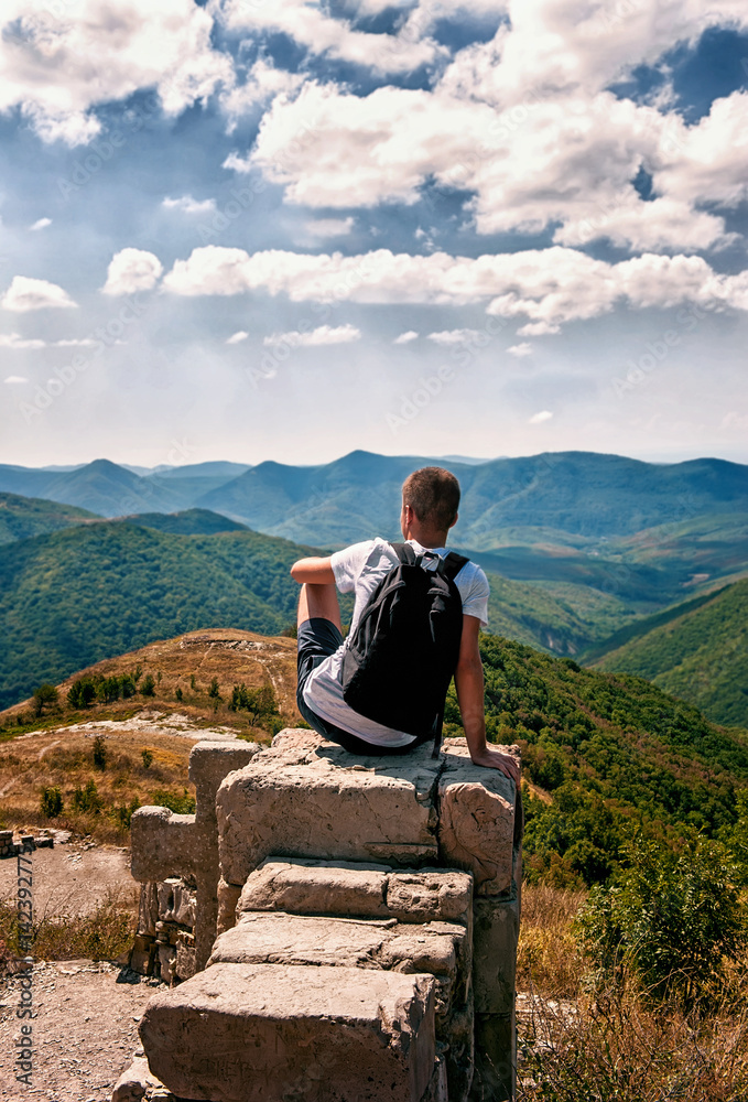 Fototapeta premium silhouette of man with backpack sitting with his back to the photographer on the big rock and looking at the panorama of mountains , sky and clouds