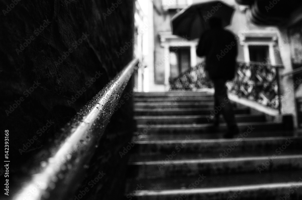 Chrome handrail with rain drops on a narrow street in Venice  while a man silhouette is walking the stairs with an umbrella