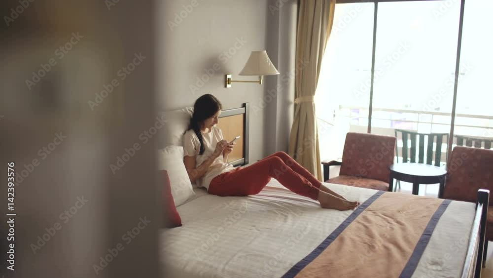 Pretty blonde woman lying in bed in hotel room and use smartphone and smiling Pretty blonde woman lying in bed in hotel room and use smartphone and smiling