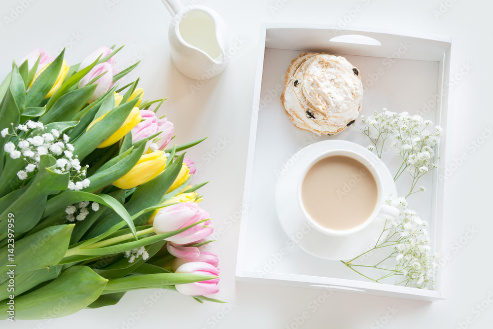 Morning breakfast in spring with a cup of black coffee with milk and pastries in the pastel colors, a bouquet of fresh yellow and pink tulips on a white background. Top view. Morning breakfast in spring with a cup of black coffee with milk and pastries in the pastel colors, a bouquet of fresh yellow and pink tulips on a white background. Top view.