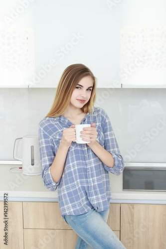 Young beautiful girl in a blue checkered shirt and jeans in the kitchen drinking tea or coffee with a white cup. Morning. home interior.