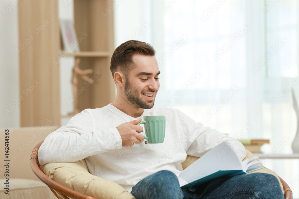 Handsome young man sitting in armchair and reading book with cup of tea at home