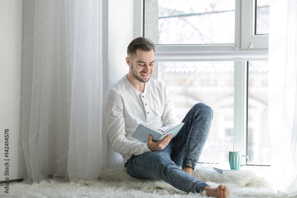 Young man sitting by the window and reading book at home