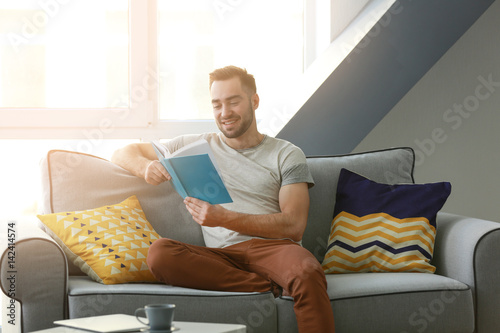 Handsome young man sitting on couch and reading book at home