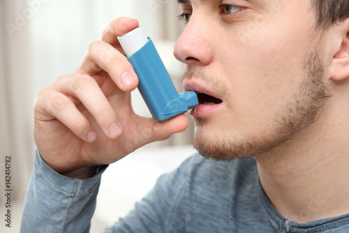 Young man using inhaler during asthmatic attack at home, closeup