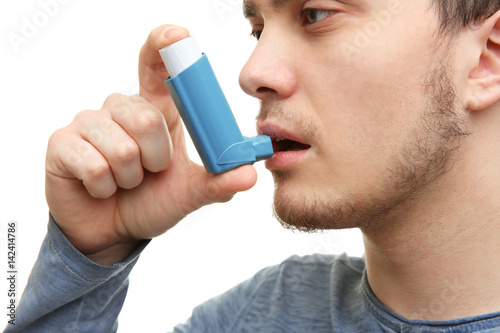 Young man using inhaler during asthmatic attack on white background, closeup