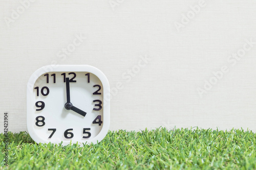 Closeup white clock for decorate in 4 o'clock on green artificial grass floor and cream wallpaper textured background with copy space photo
