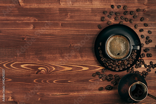 Hot coffee in black cup with crema, beans and turkish pot cezve with copy space on brown old wooden board background, top view. Rustic style.