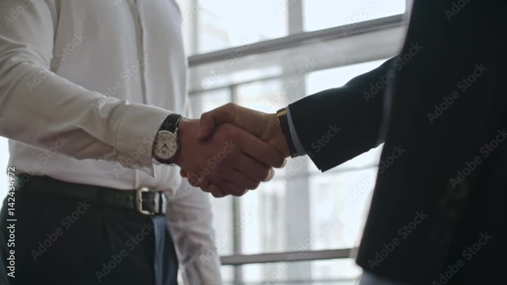 Mid-section shot with PAN of businessmen in formal wear shaking hands in agreement and discussing work before big window